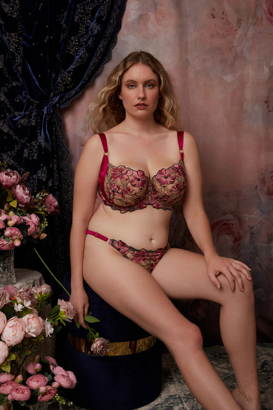 Model wearing pink and black DD+ cup bra against backdrop of pink roses and blue velvet